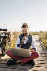 Gut gekleideter Mann sitzt auf einem Holzsteg in der Natur neben einem Fahrrad und benutzt einen Laptop - JRFF03594