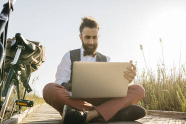 Well dressed man sitting on a wooden walkway in the countryside next to a bike using laptop - JRFF03595