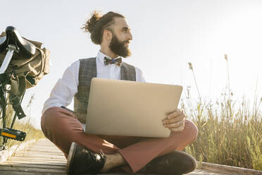 Gut gekleideter Mann, der auf einem Holzsteg in der Landschaft sitzt und mit seinem Laptop zur Seite schaut - JRFF03596