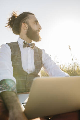 Gut gekleideter Mann, der mit einem Laptop auf dem Lande sitzt und zur Seite schaut, lizenzfreies Stockfoto