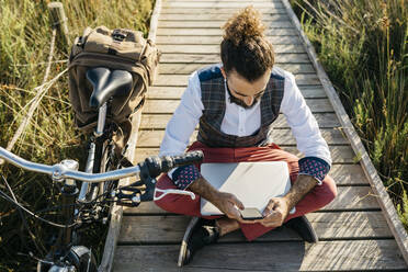 Gut gekleideter Mann sitzt auf einem Holzsteg in der Landschaft neben einem Fahrrad mit Handy und Laptop - JRFF03600