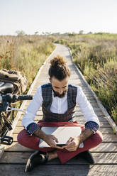 Gut gekleideter Mann sitzt auf einem Holzsteg in der Landschaft neben einem Fahrrad mit Handy und Laptop - JRFF03601