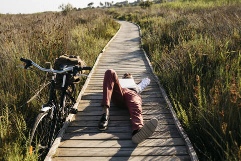 Gut gekleideter Mann mit Laptop auf einem Holzsteg in der Natur neben einem Fahrrad liegend, lizenzfreies Stockfoto