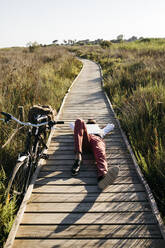 Well dressed man with laptop lying on a wooden walkway in the countryside next to a bike - JRFF03604