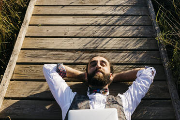 Gut gekleideter Mann mit Laptop auf einem Holzsteg in der Natur liegend - JRFF03605