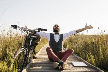 Glücklicher, gut gekleideter Mann sitzt auf einem Holzsteg in der Natur neben einem Fahrrad - JRFF03609