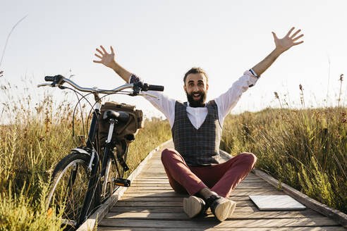 Glücklicher, gut gekleideter Mann sitzt auf einem Holzsteg in der Natur neben einem Fahrrad - JRFF03610