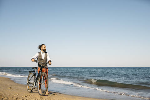 Gut gekleideter Mann, der mit seinem Fahrrad an einem Strand spazieren geht, lizenzfreies Stockfoto