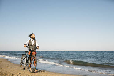 Gut gekleideter Mann, der mit seinem Fahrrad an einem Strand spazieren geht - JRFF03619