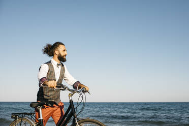 Gut gekleideter Mann, der mit seinem Fahrrad an einem Strand spazieren geht - JRFF03620