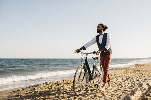 Gut gekleideter Mann, der mit seinem Fahrrad an einem Strand spazieren geht, lizenzfreies Stockfoto