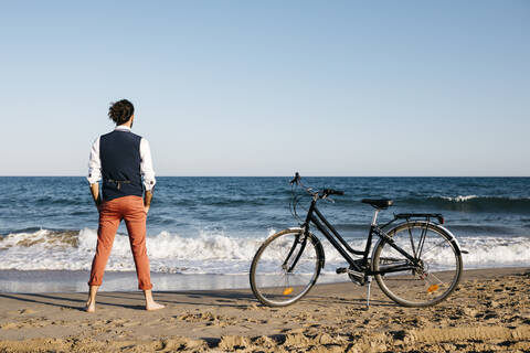 Gut gekleideter Mann mit seinem Fahrrad am Strand stehend, lizenzfreies Stockfoto