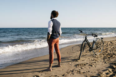 Gut gekleideter Mann mit seinem Fahrrad am Strand stehend - JRFF03623