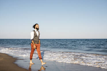 Gut gekleideter Mann, der am Strand am Wasser spazieren geht - JRFF03624