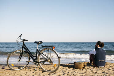 Gut gekleideter Mann mit seinem Fahrrad am Strand sitzend - JRFF03628