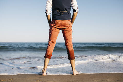 Rückansicht eines gut gekleideten Mannes, der an einem Strand am Wasser steht, lizenzfreies Stockfoto