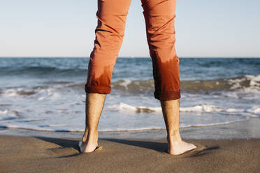 Rückansicht eines Mannes mit orangefarbener Hose, der an einem Strand am Wasser steht - JRFF03633