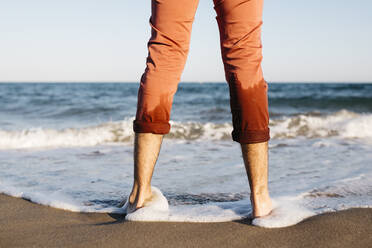 Rear view of man with orange trousers standing on a beach at water's edge - JRFF03634