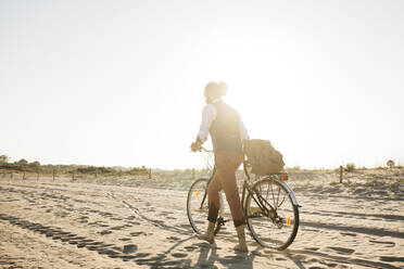 Gut gekleideter Mann, der mit seinem Fahrrad am Strand bei Sonnenuntergang spazieren geht - JRFF03635
