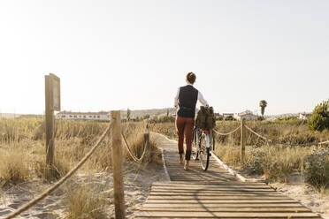 Gut gekleideter Mann, der mit seinem Fahrrad auf einer Promenade am Strand bei Sonnenuntergang spazieren geht - JRFF03636