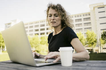 Porträt einer reifen Geschäftsfrau mit Kaffee zum Mitnehmen, die im Freien am Laptop arbeitet - KNSF06196