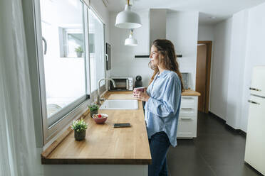 Junge Frau im Pyjama in der Küche zu Hause, die aus dem Fenster schaut - KIJF02531