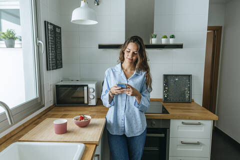 Young woman wearing pyjama in kitchen at home using cell phone stock photo