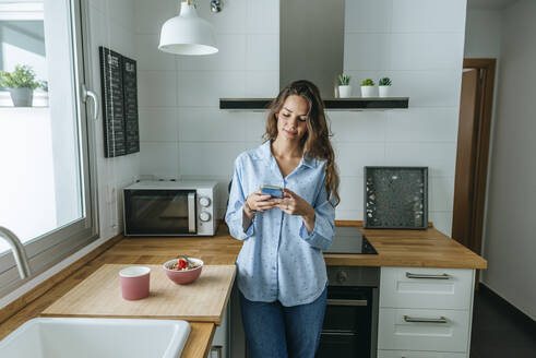 Junge Frau im Pyjama in der Küche zu Hause, die ein Mobiltelefon benutzt - KIJF02532
