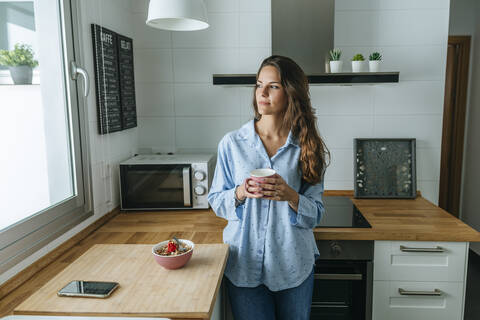 Junge Frau im Pyjama in der Küche zu Hause, die aus dem Fenster schaut, lizenzfreies Stockfoto