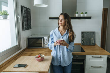 Junge Frau im Pyjama in der Küche zu Hause, die aus dem Fenster schaut - KIJF02533