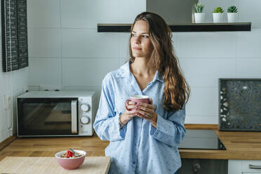 Porträt einer jungen Frau im Pyjama in der Küche zu Hause mit einer Tasse Kaffee in der Hand - KIJF02534