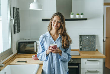 Porträt einer lächelnden jungen Frau mit einer Tasse Kaffee im Pyjama in der Küche zu Hause - KIJF02535