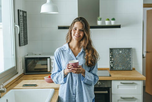Portrait of smiling young woman with cup of coffee wearing pyjama in kitchen at home - KIJF02535