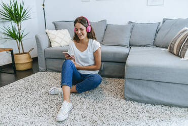 Lachende junge Frau, die im Wohnzimmer auf dem Boden sitzt und mit ihrem Mobiltelefon Musik hört - KIJF02544