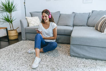 Laughing young woman sitting on floor in living room listening to music with cell phone - KIJF02544