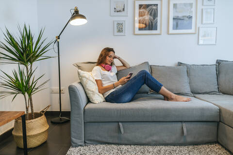 Junge Frau auf dem Sofa mit Kopfhörern und Mobiltelefon, lizenzfreies Stockfoto