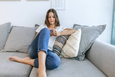 Junge Frau sitzt auf dem Sofa und benutzt ein Mobiltelefon - KIJF02551