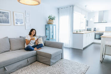 Young woman sitting on sofa with juice using cell phone - KIJF02555