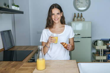 Porträt einer lächelnden jungen Frau beim Frühstück mit Saft und Croissant in der Küche - KIJF02560