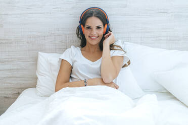 Porträt einer lächelnden jungen Frau, die mit Kopfhörern im Bett liegt - KIJF02563