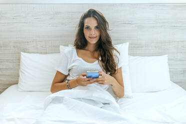 Portrait of young woman with cell phone in bed at home - KIJF02571