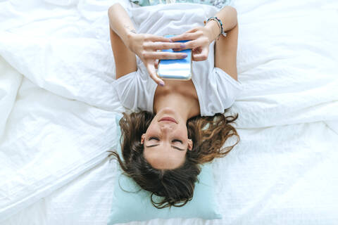 Draufsicht auf eine junge Frau, die im Bett liegt und ein Mobiltelefon benutzt, lizenzfreies Stockfoto