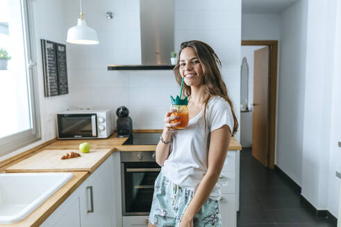Porträt einer lächelnden jungen Frau bei einem Drink in der Küche - KIJF02580