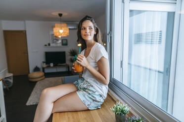 Smiling young woman sitting on kitchen counter with a drink - KIJF02582