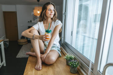 Lächelnde junge Frau sitzt mit einem Getränk am Küchentisch und schaut aus dem Fenster - KIJF02585