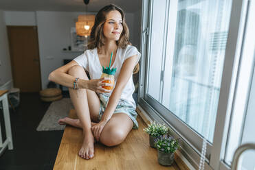 Smiling young woman sitting on kitchen counter with a drink looking out of window - KIJF02585