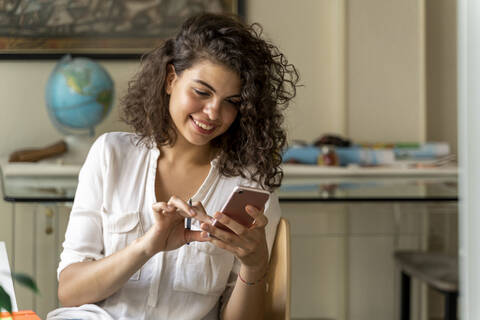 Lächelnde junge Frau, die am Schreibtisch zu Hause ein Handy benutzt, lizenzfreies Stockfoto