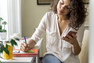 Junge Frau benutzt ein Handy und macht sich Notizen am Schreibtisch zu Hause - AFVF03724