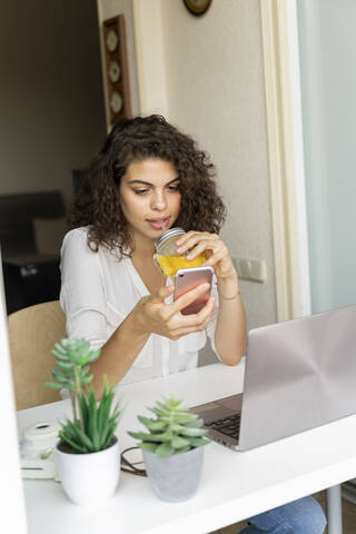 Young woman using cell phone and drinking orange juice at desk at home stock photo