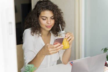 Young woman using cell phone and drinking orange juice at desk at home - AFVF03738